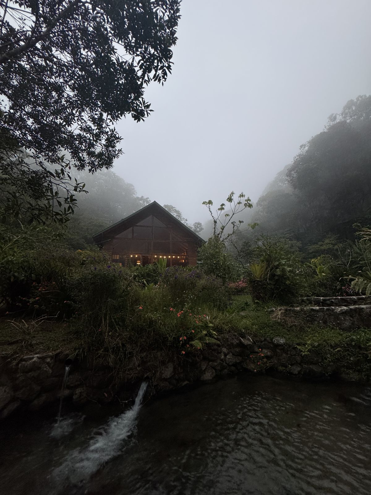 The land in cloud forest mist, a timber pavilion lit from within, water in the foreground.