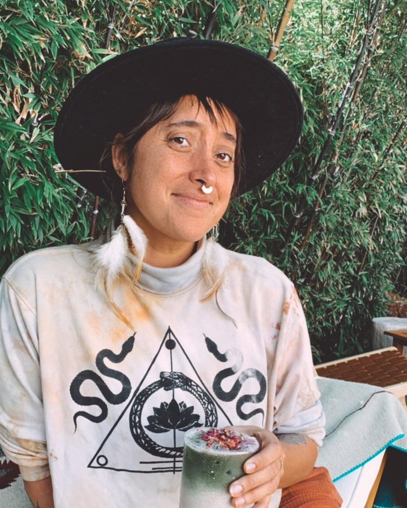 Wren, seated outdoors in a wide-brimmed hat, holding a green drink, bamboo behind her
